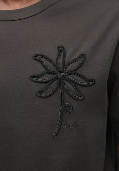 Tricou negru din bumbac cu un design floral texturat și brodat în negru pe zona stângă a pieptului, având detalii de cusătură vizibile.