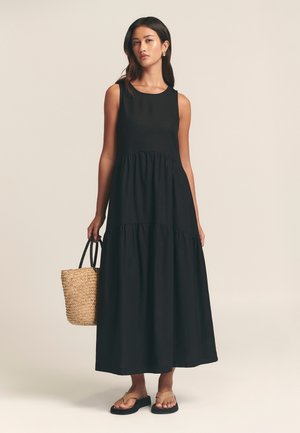 Black sleeveless maxi dress with a tiered hem, made of soft fabric. Paired with a woven straw tote bag and flat sandals.