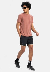 Vīrietis staigā, valkājot brūnu T-kreklu, melnas šortus ar bēšām malām, melnas zeķes, melnus sporta apavus un lielas melnas saulesbrilles uz baltas fona.