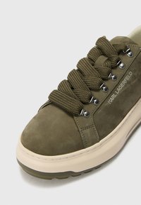 Olive green suede sneaker with thick textured laces, silver eyelets, contrasting cream rubber sole, and embossed logo on side.