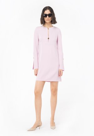 Mujer con un vestido mini de manga larga en rosa claro con botones dorados, tacones de kitten beige y gafas de sol negras, de pie contra un fondo blanco.