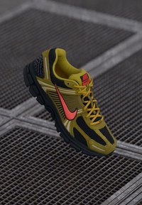 Žlté a čierne bežecké tenisky Nike s oranžovým logom a šnúrkami ležiace na povrchu kovovej mriežky.