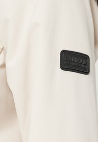 Μανίκι από υφασμάτινο σακάκι κρεμ υφής με μαύρο ορθογώνιο λογότυπο Barbour International ραμμένο κοντά στον ώμο.