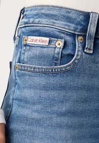 Svijetloplave traperice s prednjim džepom, zlatnim detaljima i crveno-bijelom oznakom "Calvin Klein" ušivenom iznad.