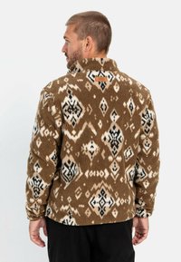Braune Fleecejacke mit einem strukturierten Muster aus cremefarbenen und schwarzen geometrischen Formen; verfügt über einen hohen Kragen und ein Lederetikett auf der Rückseite.