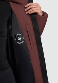 Burgunderfarbenes und schwarzes isoliertes Kleidungsstück mit einer glatten Außenfläche, ausgestattet mit einem Logo, einer Innentasche und einem eleganten, modernen Finish.