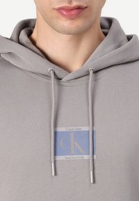 Šedá mikina s kapucňou a šnúrkami. Predná časť obsahuje nášivku s logom "Calvin Klein," "cK," a "New York City" v modrej a šedej farbe.