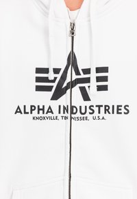 Biely zipsový mikina s čiernym logom obsahujúcim horizontálne pruhy a text, ktorý uvádza „ALPHA INDUSTRIES, KNOXVILLE, TENNESSEE, U.S.A.“