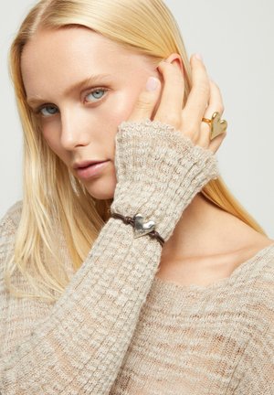 Mujer rubia vistiendo un suéter de punto beige, mostrando un anillo dorado en forma de corazón y una pulsera de plata con forma de corazón en su muñeca.