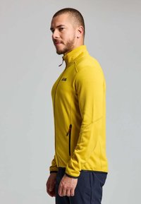 Man wearing a bright yellow zip-up jacket with black zipper details, standing in profile against a plain light gray background.