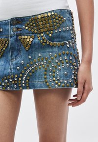 Τζιν μίνι φούστα με χρυσά και ασημένια καρφιά σε διάφορα μεγέθη, με διακοσμητικές τσέπες και ελαφρώς φθαρμένο τελείωμα.