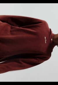 Personne portant un shirt à manches longues bordeaux avec "Bobo Choses" brodé sur la poitrine, se tenant devant un fond clair uni.