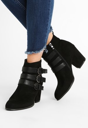Bottines en daim noir à la cheville avec trois sangles en cuir à boucle et fermeture éclair latérale, portées avec un jean skinny bleu à ourlet effiloché.