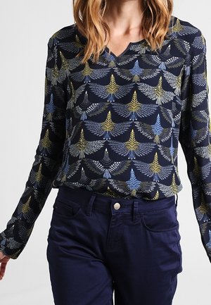 Blouse marine à manches longues avec des motifs d’ailes d’oiseaux en bleu, jaune et blanc, associée à un pantalon bleu foncé, portée par une personne aux cheveux mi-longs.