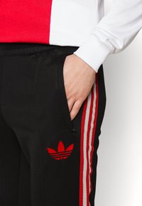 Black athletic pants with red Adidas logo, side pocket, and red and white striped accents along the sides; smooth texture and fitted design.