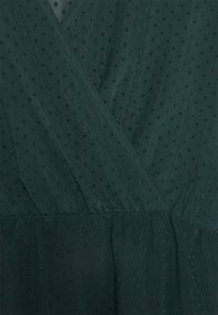 Dunkelgrünes Wickelkleid aus strukturiertem Stoff, mit kleinen schwarzen Pünktchen, einem tiefen V-Ausschnitt und einem gerafften Taillendetail.