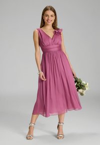 Robe à encolure en V, longueur genou en tissu rose clair et doux. Dotée d'un accent floral sur l'épaule gauche et d'une taille ajustée. Chaussures argentées.