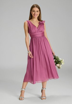 Kleid mit V-Ausschnitt in knielanger Länge aus einem sanften, hellrosa Stoff. Verfügt über eine Blumenapplikation an der linken Schulter und einen taillierten Bund. Silberne Schuhe.