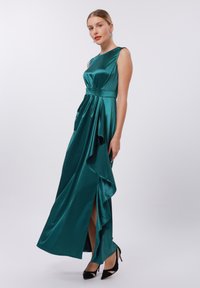 Tealfärgad satin aftonklänning med figursydd överdel, draperad kjol, hög slits och asymmetrisk fåll, kombinerad med svarta spetsiga klackar.