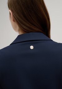 Marine shirt met een kraag, voorzien van een zilveren ronde knop op de rug. Gladde textuur, minimalistisch ontwerp.
