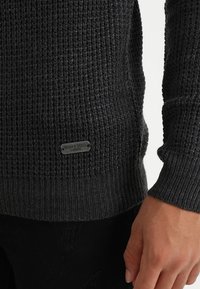 Mörkgrå stickad tröja med ett strukturerat mönster och en metallskylt i botten med texten "BRAVE SOUL LONDON" graverad.