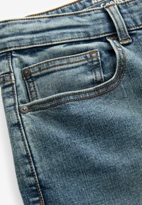 Jeans din denim de culoare albastră deschisă, cu o textură uzată. Au două buzunare cu obturatoare metalice și cusături contrastante de culoare maro pe cusături.