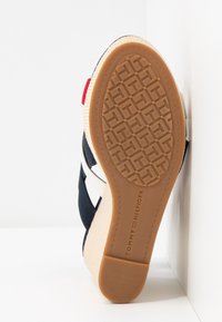 Sandale compensée avec une tige en tissu bleu marine, semelle de style espadrille beige et semelle extérieure en caoutchouc texturé avec un motif géométrique. Détail en accent rouge.