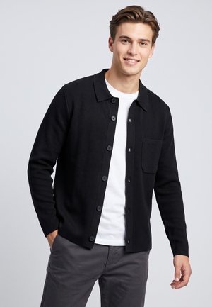 Junger Mann trägt eine schwarze Strickjacke mit Knöpfen über einem weißen T-Shirt und grauen Hosen, steht mit einer Hand in der Tasche und lächelt in die Kamera.