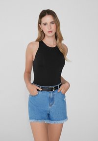 Joven mujer con cabello largo y rubio, vistiendo una camiseta sin mangas negra, pantalones cortos de mezclilla azules con bordes deshilachados y un cinturón negro, posando con las manos en los bolsillos.