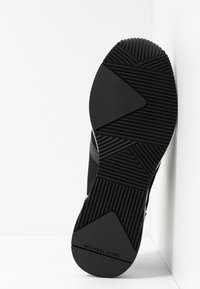 Černá podešev tenisky z texturované gumy s geometrickým vzorem, značkou a hladkým okrajem. Navržena pro lepší přilnavost a odolnost.