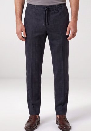 Man draagt donkergrijze geruite broek met trekkoord in de taille en bruine leren loafers, staat op een witte achtergrond.
