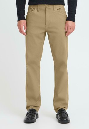 Pantalon en coton beige avec une coupe droite, doté de deux poches latérales et d'une fermeture à bouton sur le devant, associé à des chaussures noires.