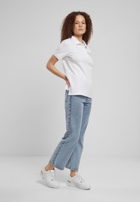 Wit poloshirt van lichtgewicht stof, gecombineerd met lichtblauwe jeans met rechte pijpen en witte sneakers. Krullend haar, handen in de zij, zijaanzicht.