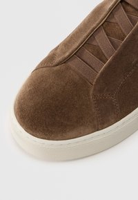 Καφέ σουέντ αθλητικό παπούτσι με στρογγυλεμένη μύτη, υφασμάτινες ελαστικές λεπτομέρειες και λευκή καουτσούκ σόλα με υφή γι α καλύτερο κράτημα.