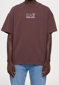 Bordowa, luźna bawełniana koszulka z krótkim rękawem, z białym logo „EA7 Emporio Armani” na piersi. Casualowy krój i miękka tekstura.
