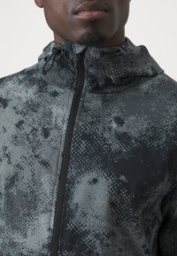 Veste à motif noir et gris avec fermeture éclair intégrale, capuche ajustable et tissu texturé avec un léger éclat. Logo Nike sur la poitrine.