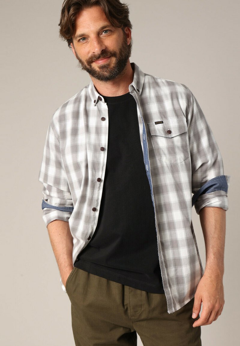 Grijs en wit geruit overhemd met knopen in bruin, opgerolde mouwen en een borstzak, gedragen over een zwart shirt en olijfgroene broek.