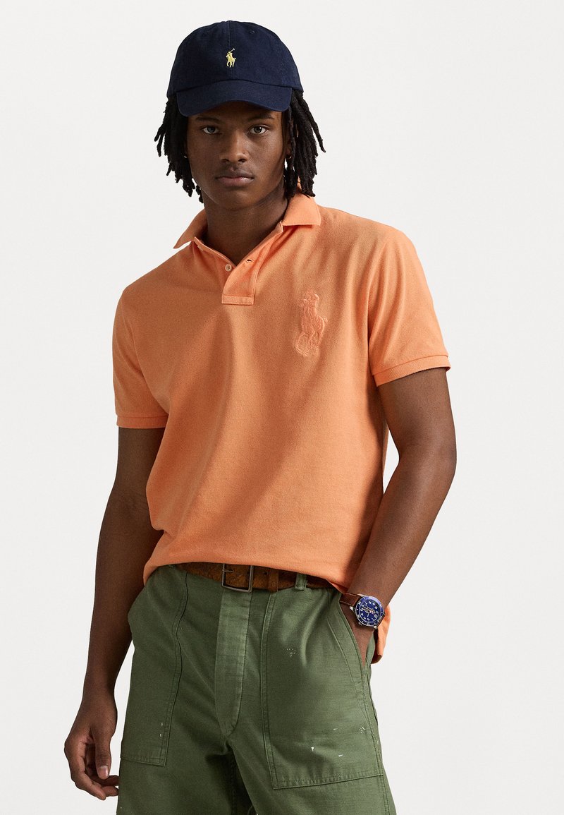 Oranje katoenen polo met een gestructureerde afwerking en geborduurd logo op de borst, gecombineerd met groene cargobroeken en een marineblauwe pet.
