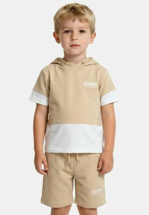 Garçon blond portant un sweat à capuche à manches courtes beige et blanc ainsi qu'un short beige assorti avec des logos de marque, debout devant un fond blanc uni.