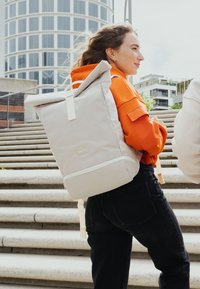 Fiatal nő narancssárga kapucnis felsőt és fekete farmert visel, miközben egy nagy bézs hátizsákot cipel a városban található kültéri lépcsőkön.