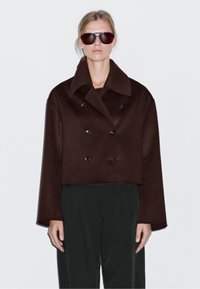 Brązowy krótki płaszcz podwójnie zapinany na duże guziki, z szerokim kołnierzem, zestawiony z luźnymi ciemnymi spodniami. Model nosi także ciemne okulary przeciwsłoneczne.