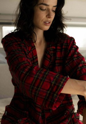 Kobieta z ciemnymi włosami w czerwonej kraciastej koszuli patrzy w dół, sięgając przed siebie w miękko oświetlonym pokoju.