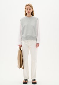 Felpa grigia con orlo a costine, maniche lunghe bianche, jeans color crema e sandali neri. Sostenendo una borsa a tracolla color sabbia, in piedi su uno sfondo bianco.