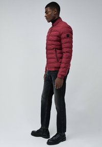 Chaqueta acolchada roja con cuello alto, bolsillos con cremallera y textura acolchada. Combinada con jeans oscuros y botas negras con cordones.