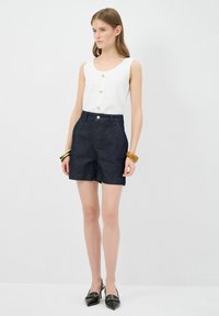 Vit ärmlös topp med horisontella ränder och guldknappar, kombinerad med mörka denimshorts med hög midja och knappstängning.