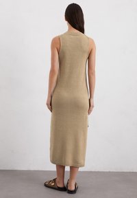 Vestito senza maniche in maglia beige, caratterizzato da un colletto alto, bordi a coste e lunghezza fino al polpaccio con spacchi laterali. Indossato con sandali piatti.