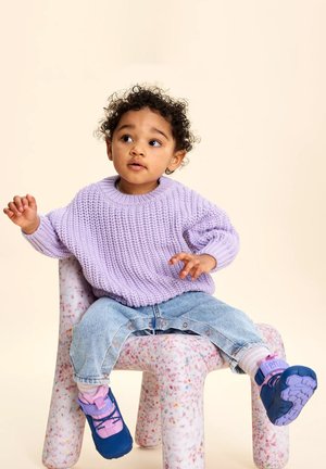 Kleinkind mit lockigem Haar, das einen lila Pullover und blaue Schuhe trägt, sitzt auf einem rosa-weiß gesprenkelten Stuhl und blickt leicht zur Seite.