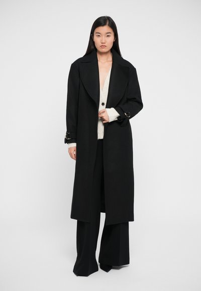 Palton lung negru cu rever larg, accente aurii la mansete, purtat peste un cardigan gri deschis și pantaloni negri evazați. Textură de țesătură netedă.