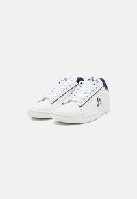 Baskets blanches avec une finition en cuir lisse, accents contrastants bleu marine, lacets plats, et un logo sur le côté et la languette. Semelle intérieure rembourrée visible.