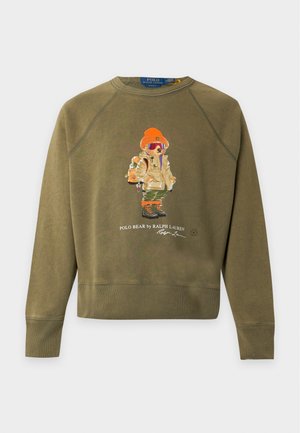 Giovane uomo che indossa una felpa verde oliva con una grafica dell’orso, jeans larghi azzurri chiari e mocassini beige, in piedi davanti a uno sfondo neutro.
